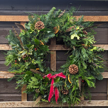 Load image into Gallery viewer, Christmas wreath with greenery, pine cones, and a red ribbon on a wooden background